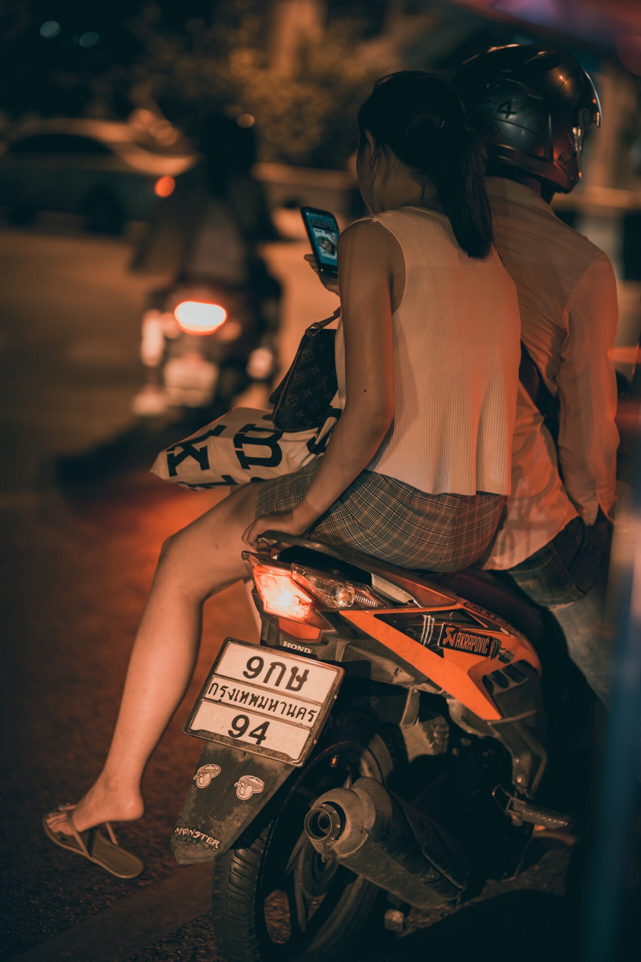 In the glow of the streetlights, a passenger on a motorbike checks her phone, highlighting the interplay of connectivity and mobility in the urban night.