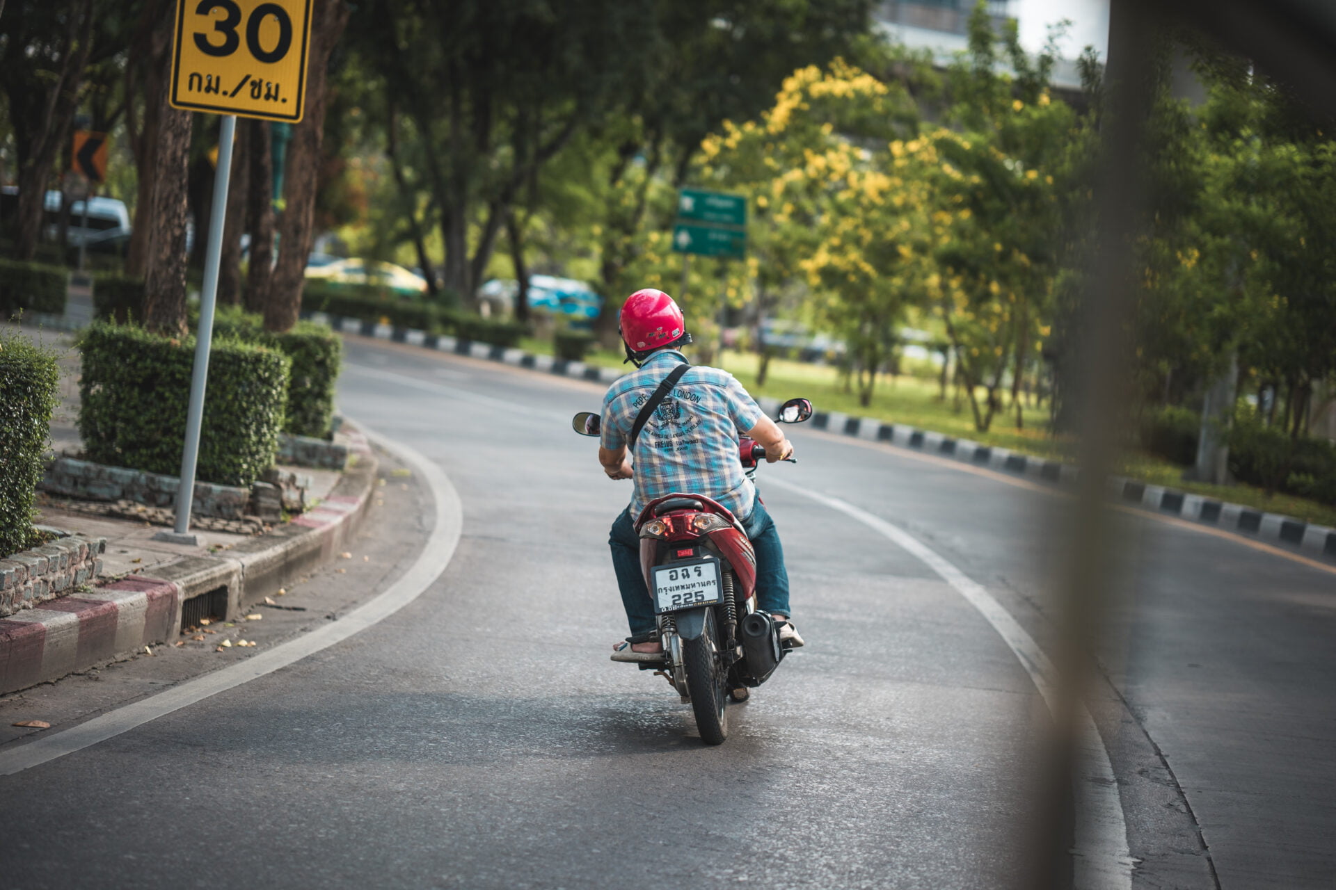 A lone rider in a plaid shirt and vibrant pink helmet maneuvers a red scooter on an empty city road, flanked by lush greenery and yellow flowering trees. A 30 km/h speed limit sign stands by, hinting at a serene, suburban locale. The scene is captured from a perspective that gives a sense of motion, with the scooter slightly off-center, suggesting a moment of peaceful transit in the everyday hustle.