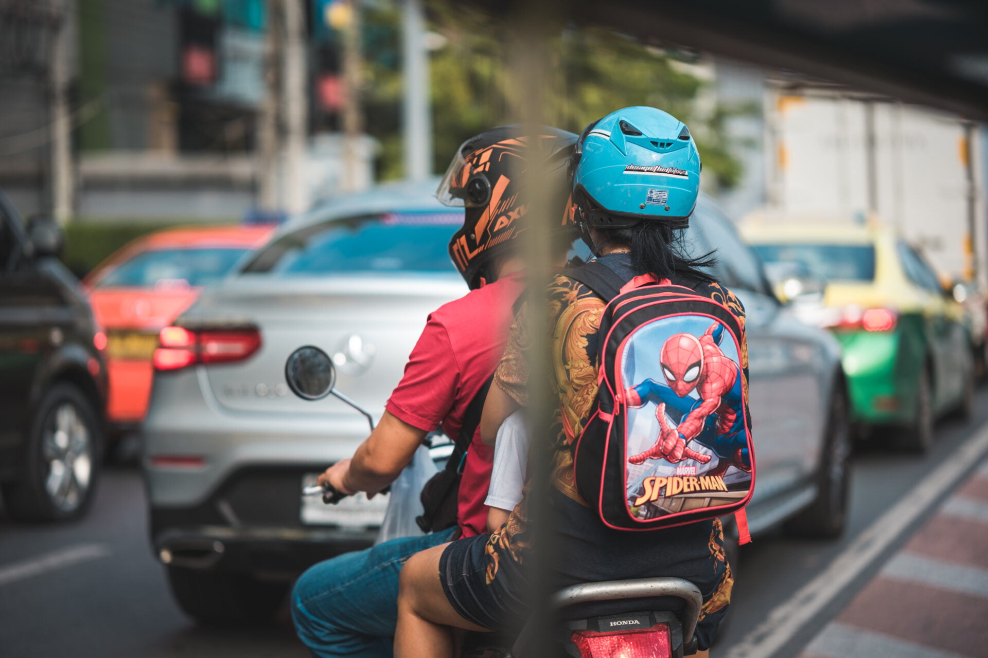 A motorcyclist wearing a blue helmet and a passenger with a Spider-Man backpack riding through congested city traffic, with cars and a taxi visible in the background.