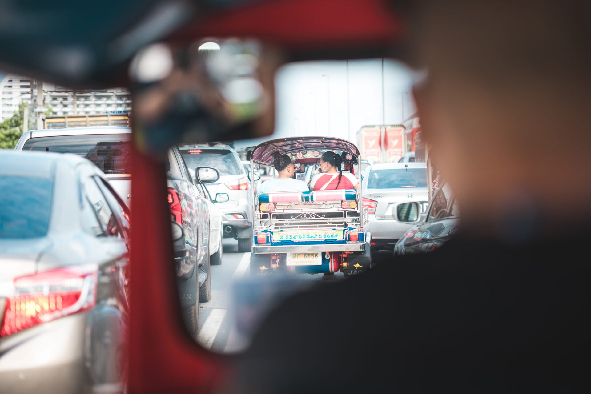 View through a vehicle's interior showing a busy street scene with cars and a colorfully decorated auto-rickshaw carrying passengers, in a conversation, amidst a traffic jam.