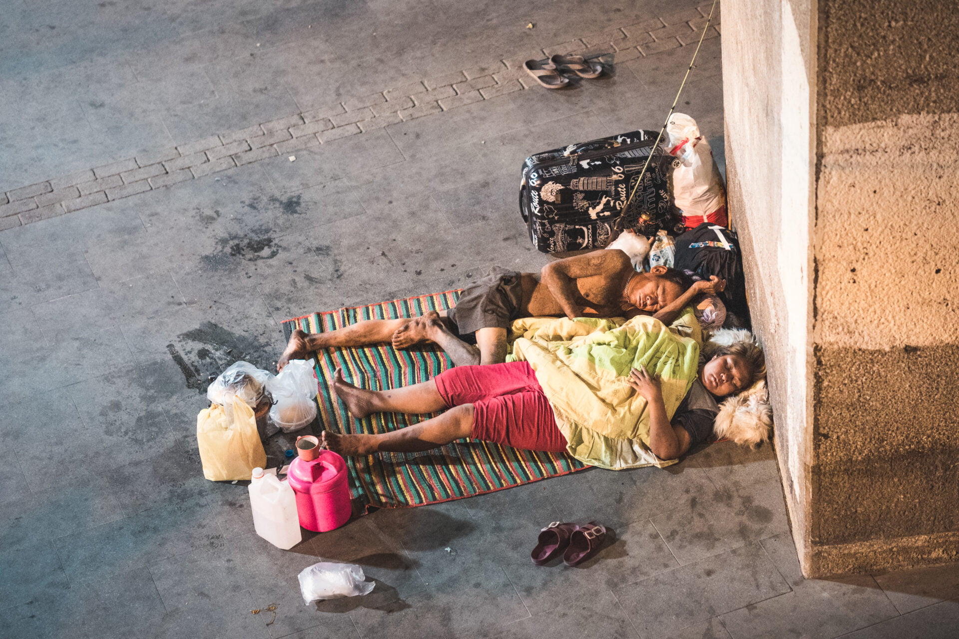 An overhead view of two people sleeping on a colorful mat on the pavement, surrounded by personal belongings and plastic containers, with a pair of flip-flops placed neatly to the side.