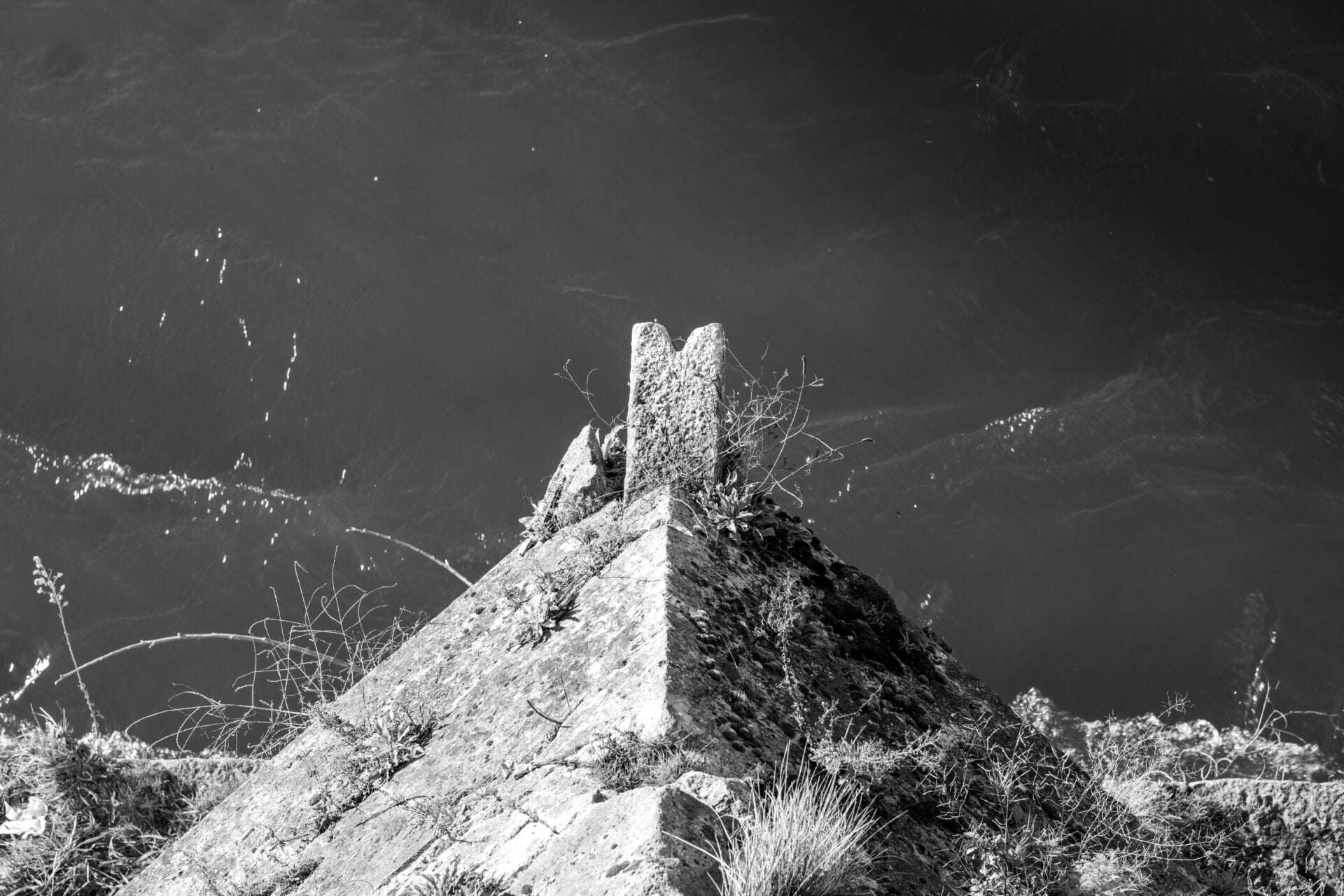 A black and white photo captures a pointed rock formation resembling a small mountain peak, jutting out against a backdrop of water with ripples and reflections glinting in the light.