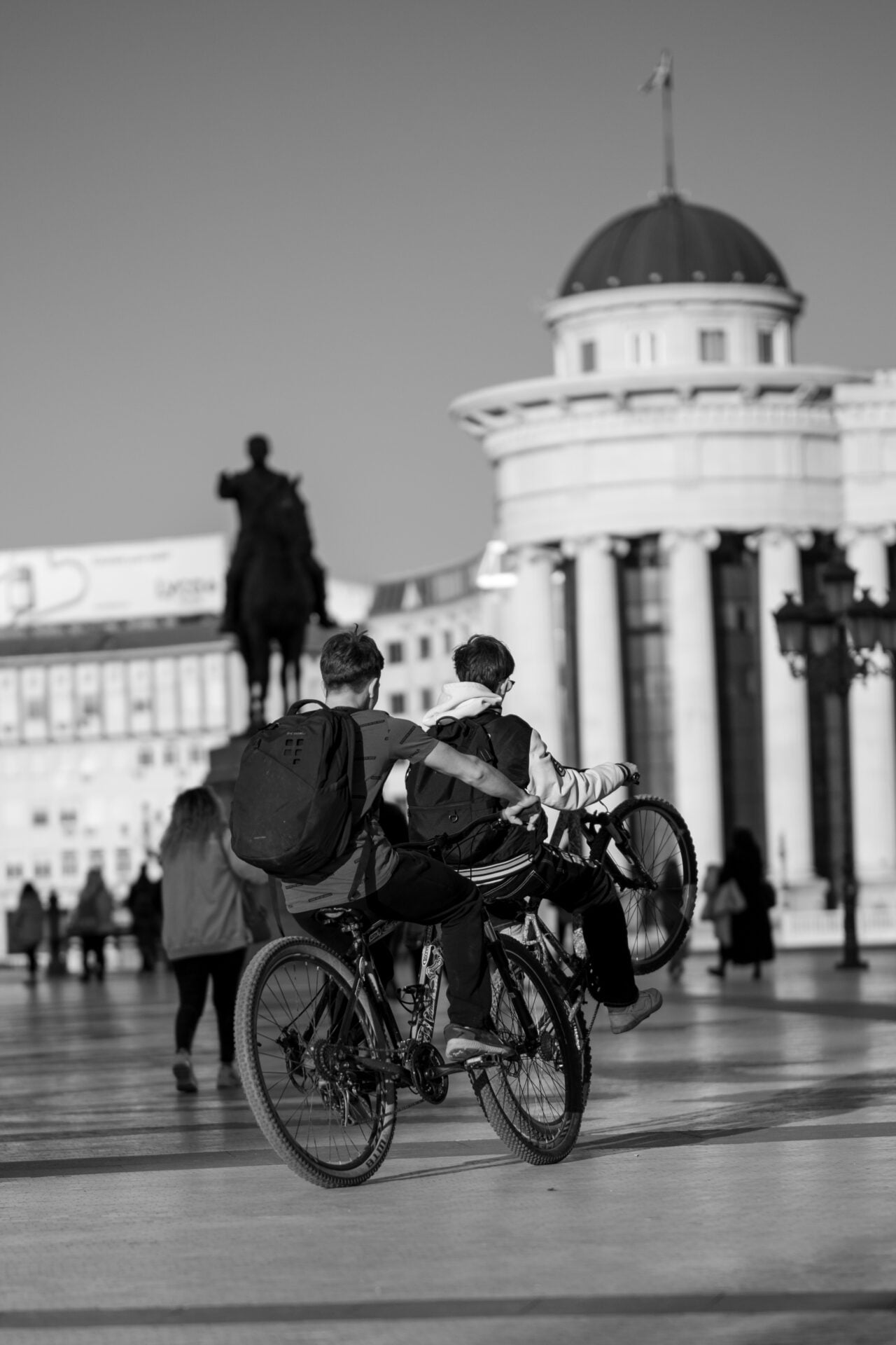 In a black and white photo, two individuals on a bicycle, sporting backpacks, navigate a bustling city square, with a statue and classical architecture framing their journey.