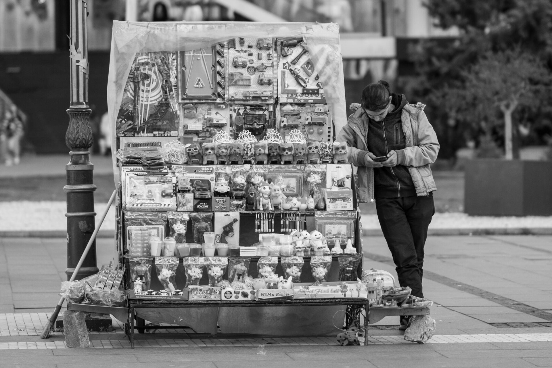 In a monochrome photo, a street vendor in a jacket leans over, focused on his phone, beside a stand displaying an array of items including toys and trinkets, set against an urban sidewalk scene.