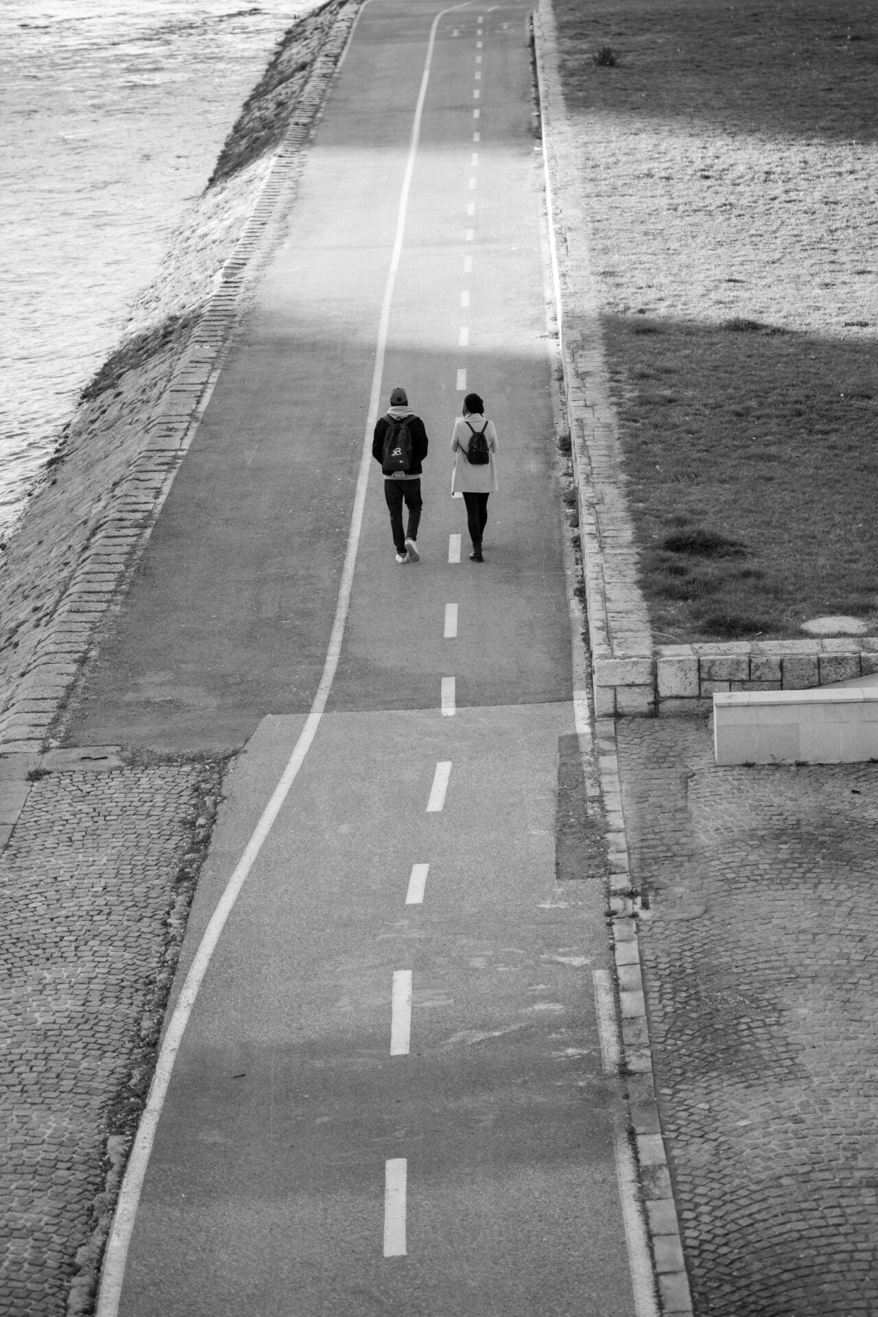 A black and white photo captures two people walking side by side along a paved path with bicycle lane markings, adjacent to a waterway, embodying a peaceful coexistence.