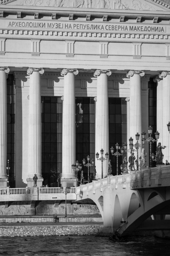 Black and white image displaying the neoclassical architecture of the Archaeological Museum of North Macedonia, with its prominent Corinthian columns and intricate frieze, juxtaposed against the flowing river and a modern bridge adorned with street lamps.