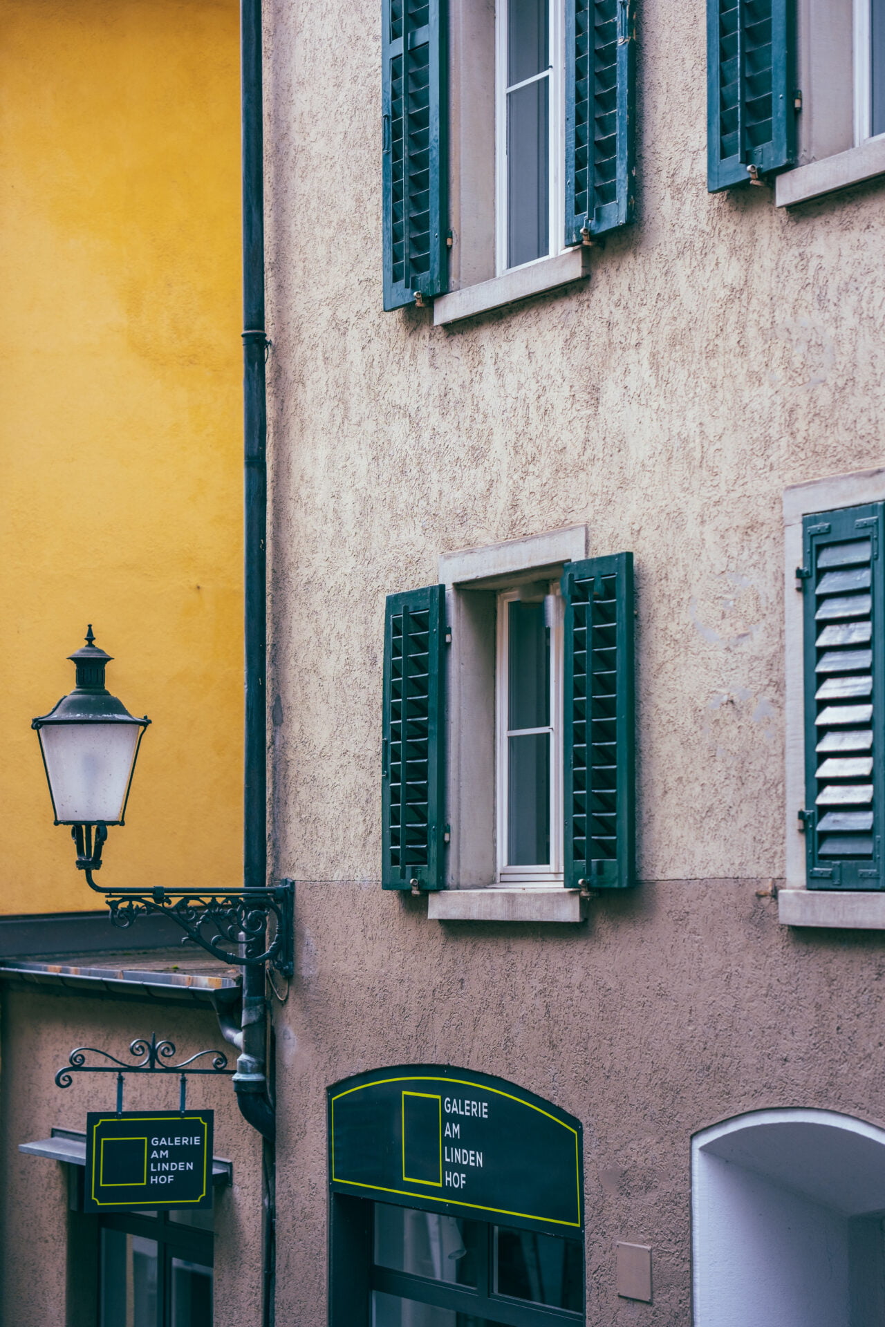 This frame is rich with urban texture: the mustard walls, the street lamp's old-timey vibe, and those shutters whispering tales of daily life. Peep the sign for "Galerie Am Lindenhof" - a hint of local art and culture.