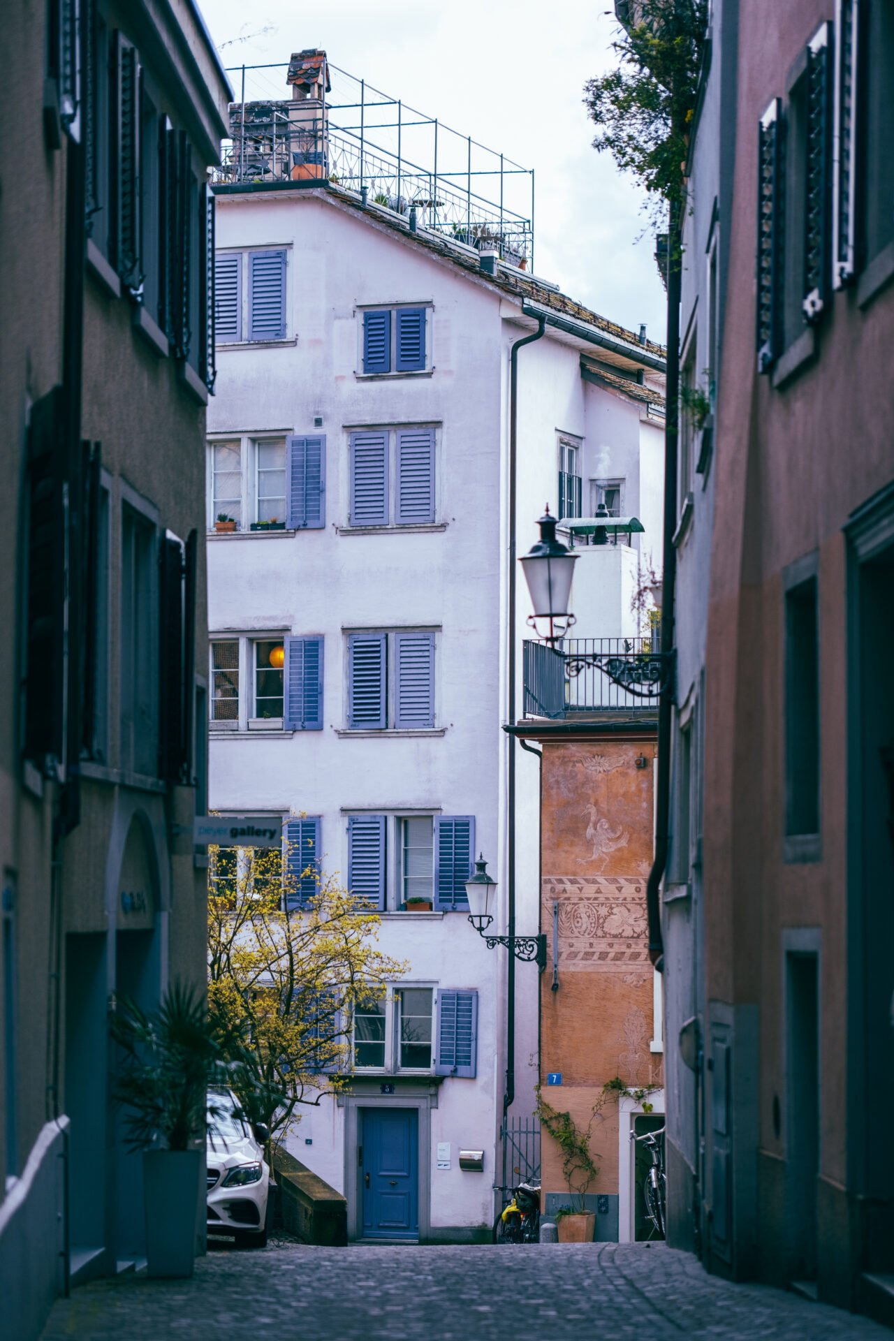 A cobblestone street carves its way between pastel buildings, their shutters a silent nod to the stories within. A lone balcony boasts greenery against the sky, while a burst of yellow from a flowering tree adds a pop of life. It's a slice of serene city living.