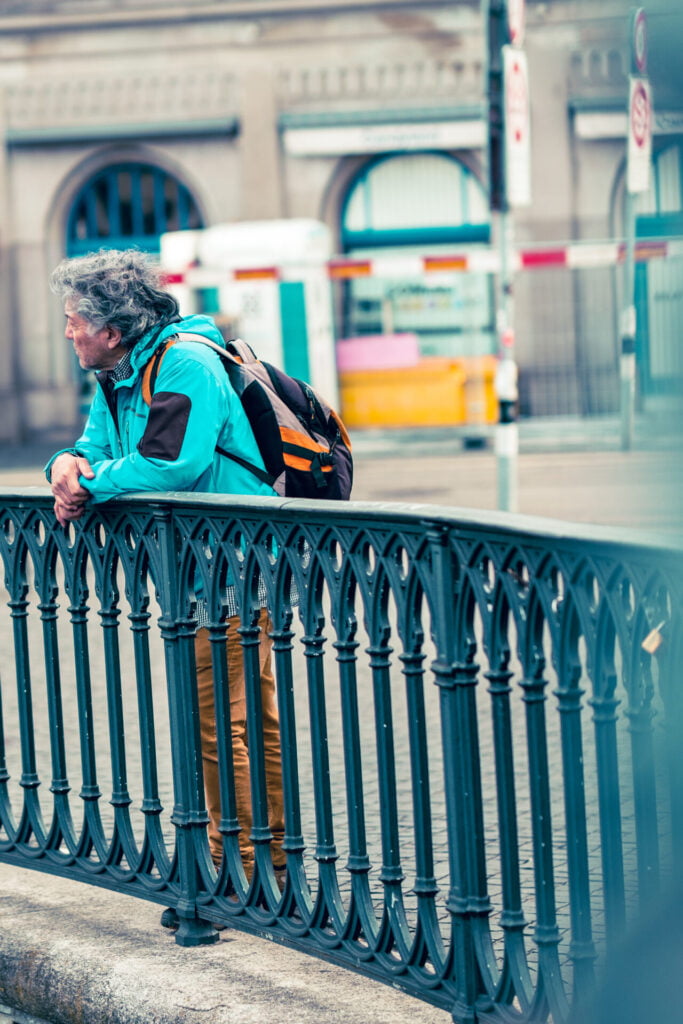 A person caught in a moment of rest, gaze cast out perhaps in thought or observation. They blend into the urban tapestry, a figure among the flow of city life, poised against the ornate railing that hints at a boundary between the bustle and a moment’s peace.