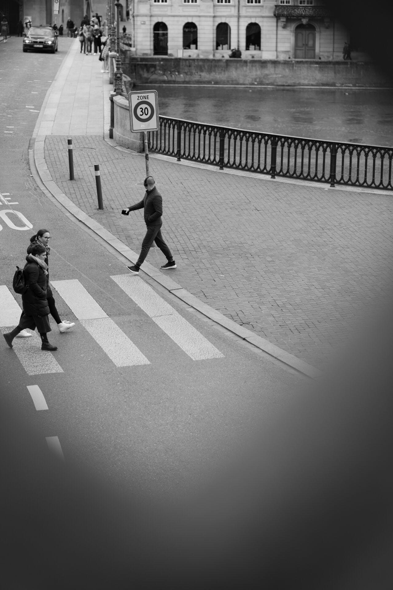 A black and white world, where pedestrians set the rhythm. One strides confidently across the crosswalk, a silent metronome against the city's tempo. Meanwhile, life flutters by — a pair of walkers in sync, edged out by the water's quiet murmur. It's urban poetry in motion.