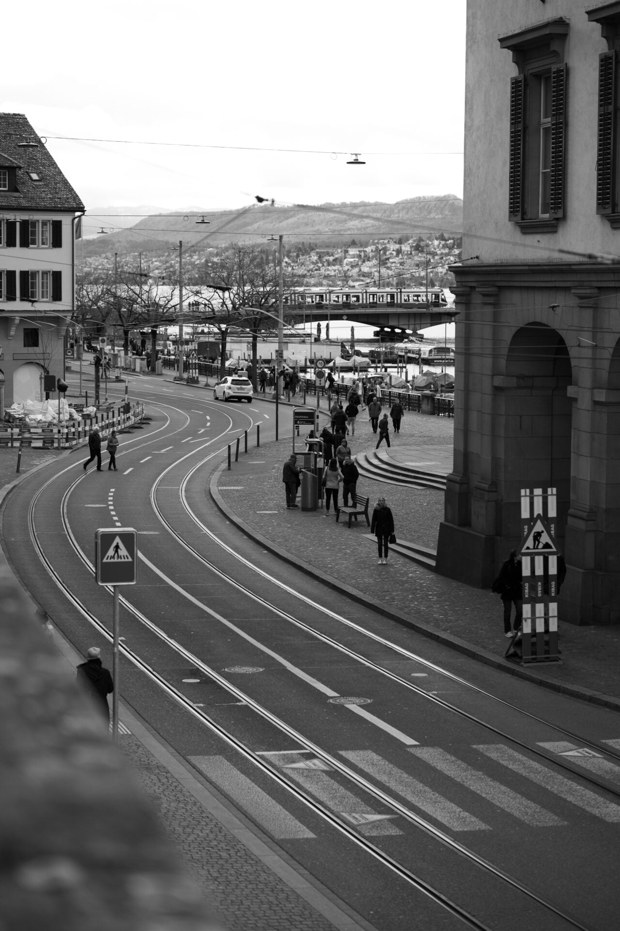 Curves and lines guide the eye, tracing paths where people and vehicles dance in a daily ballet. The scene unfolds in grayscale, inviting contemplation. It's a snapshot of routine, set against a backdrop of distant hills, whispering tales of beyond.