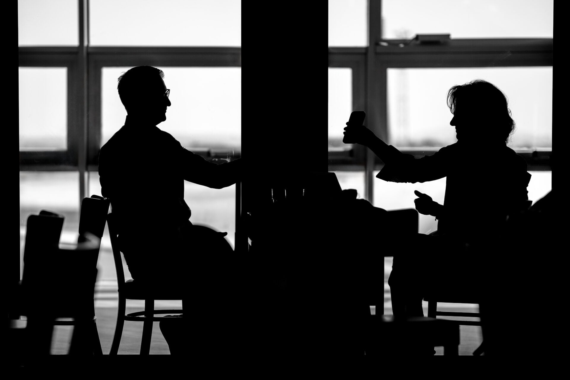 Silhouette of two people facing each other in conversation, one holding a mobile phone, backlit by large windows with a bright sky beyond.