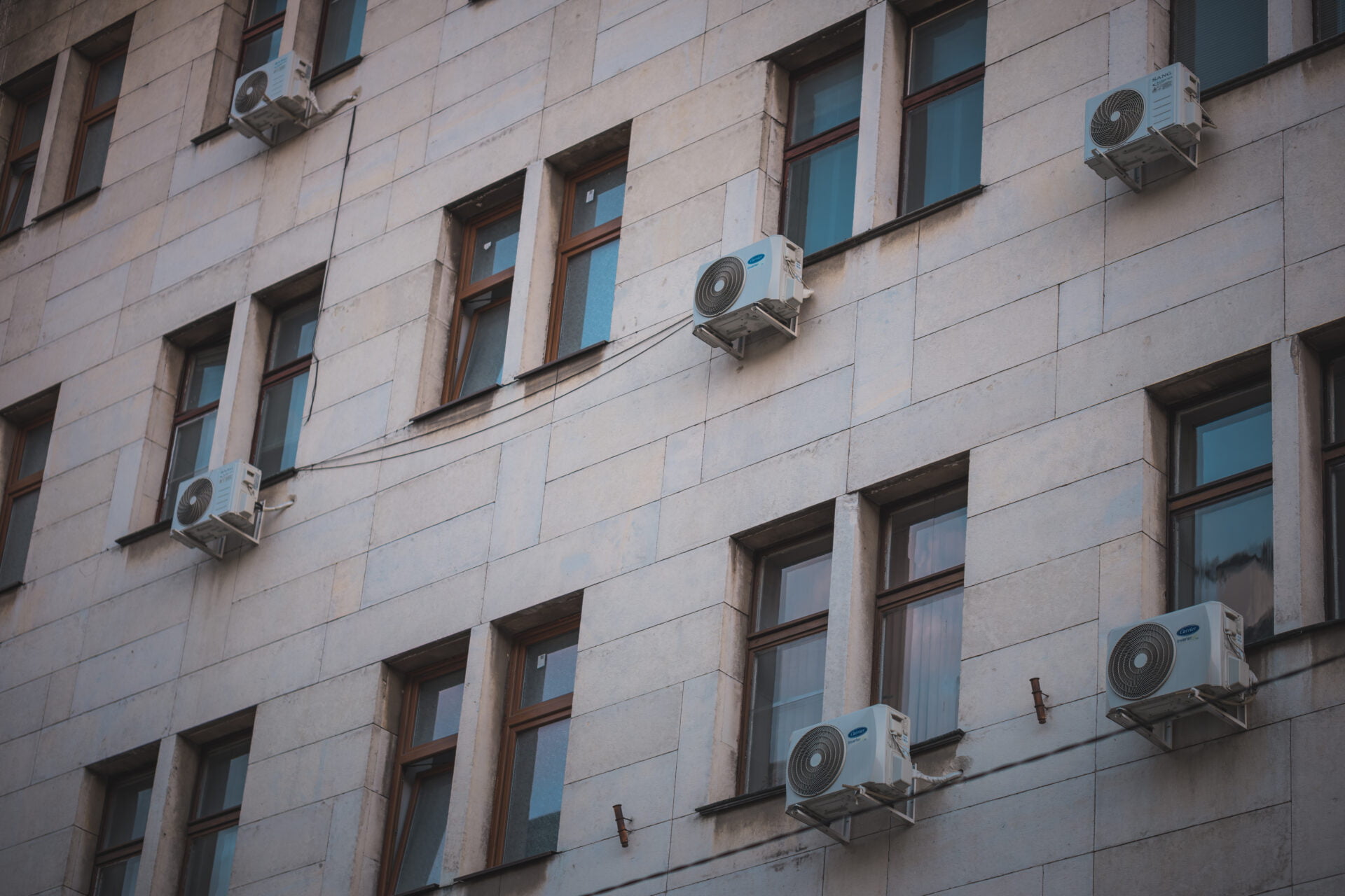 Three air conditioning units mounted on the exterior wall of a building with multiple windows.
