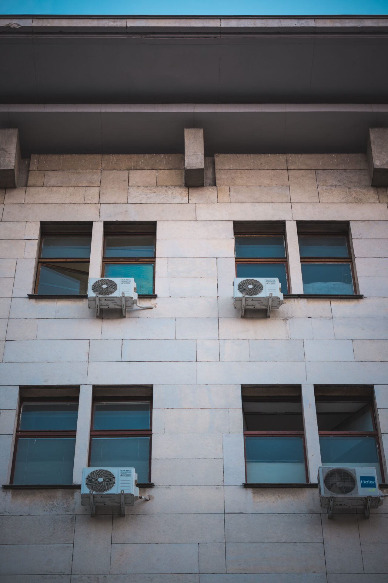 Three air conditioning units mounted below windows on a building's stone facade, showcasing a blend of functionality and symmetry.