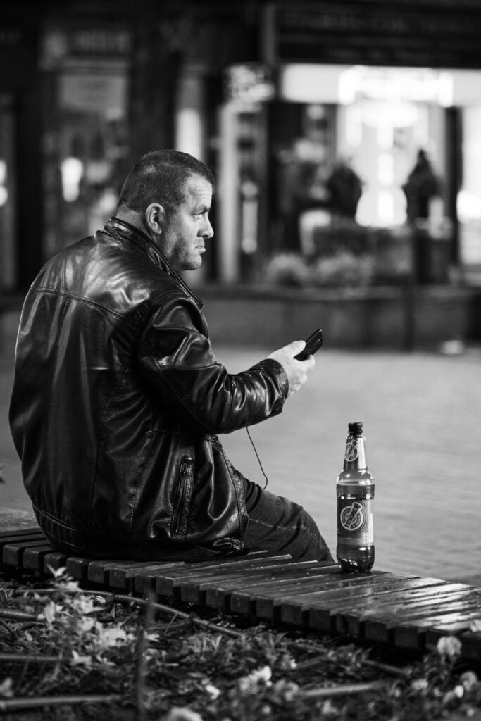In a black and white photo, a man in a leather jacket sits on a bench, focused on his smartphone, with a bottle beside him on the bench.