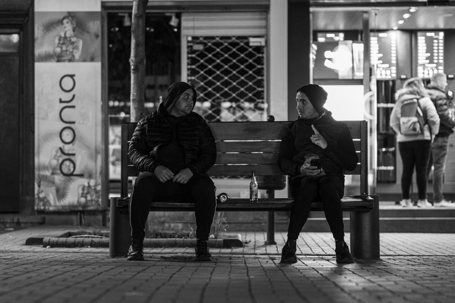 Black and white image of two people sitting on a bench at night, talking, with blurred figures in the background.
