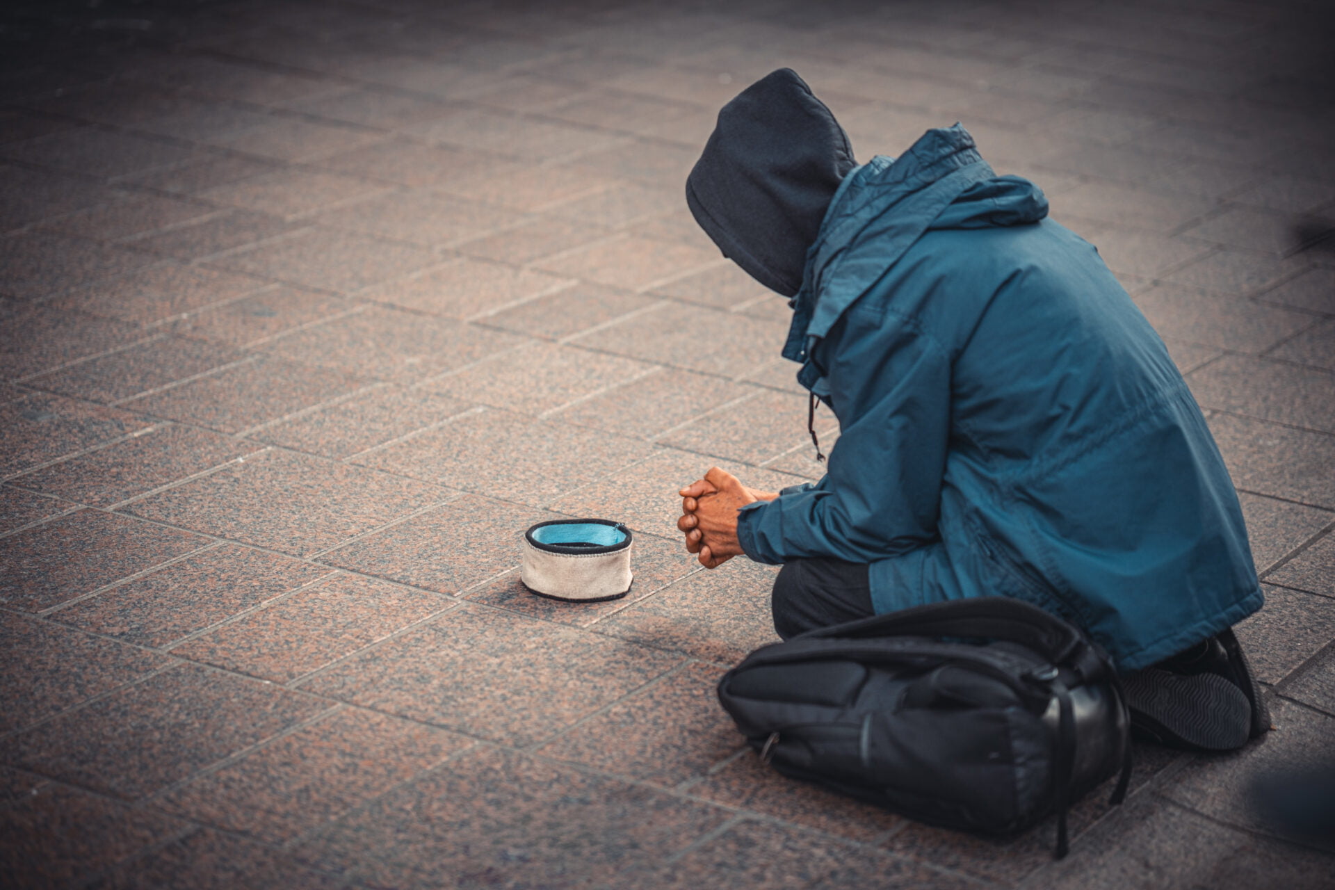 An individual in a blue jacket and hood, kneeling on the street with a hat placed in front for donations.