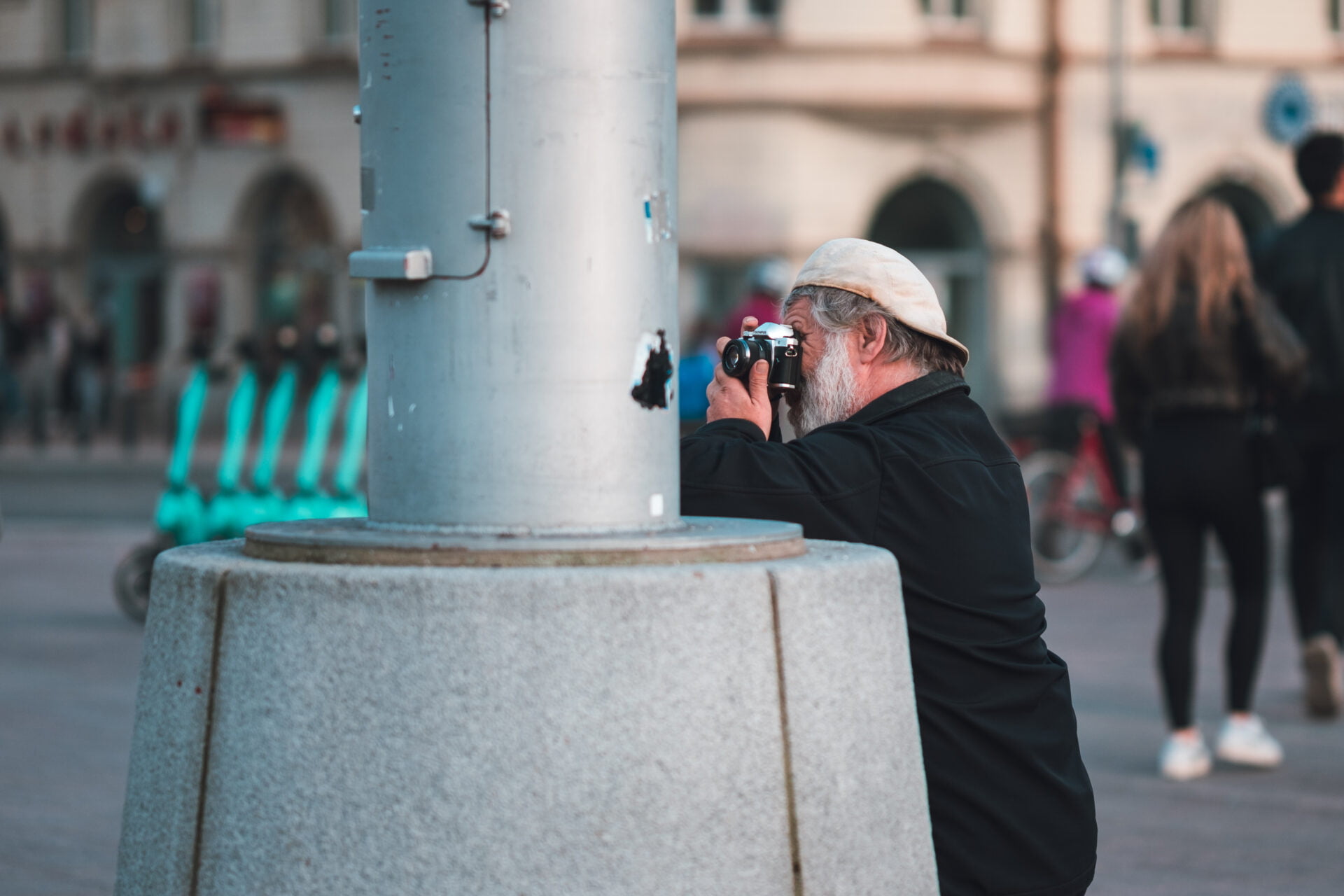 Urban artist: A seasoned photographer captures life's candid moments on bustling city streets. - Milan Ivanovic Photography A person with a cap and beard taking a photograph in a city scene, focused intently on their subject.