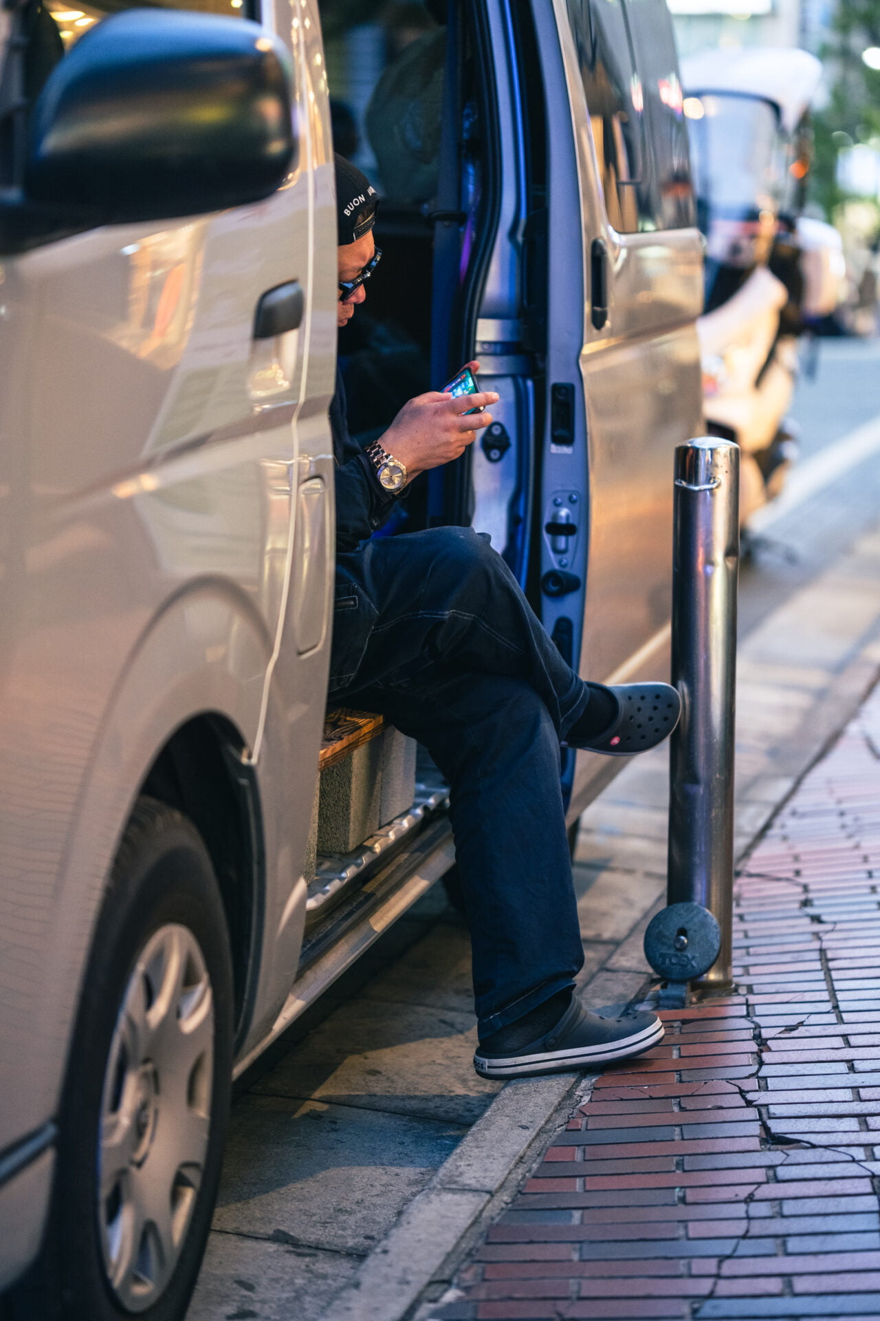 A man sits casually in the open doorway of a parked van, focused on his smartphone, his black beanie and casual attire contrasting with the sleek metallic exterior of the vehicle and the urban pavement, symbolizing a moment of stillness in the city's rhythm.